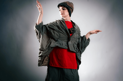 Model wearing an oversized charcoal jacket over a bright red outfit with a textured knit hat, posing to showcase an avant-garde fashion look with sculptural silhouettes.